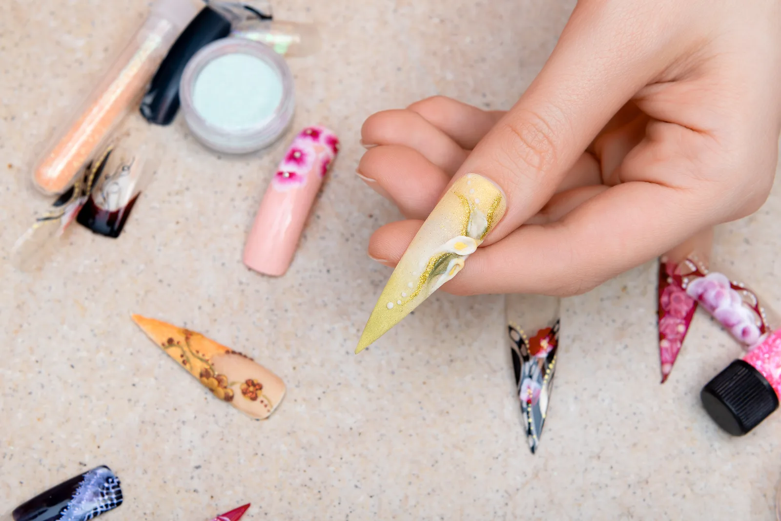 A hand holding a long stiletto nail tip with hand-painted lily nail art, surrounded by more decorated tips and pots of glitter on a speckled surface